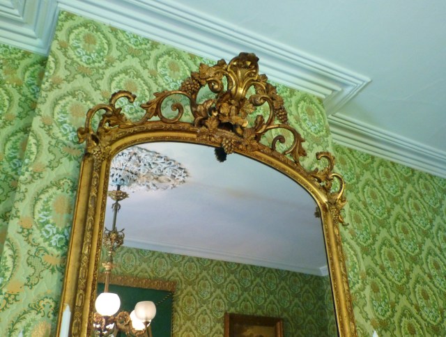 The glorious gilt mirror above the fireplace mantel goes all the way to the crown moulding.  It is adorned with ornate scrolls and sculpted grapes.  Reflected in the mirror is the equally ornate plaster ceiling medallion from which the chandelier is suspended. 