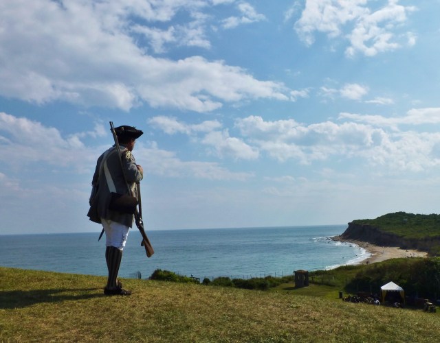 A gentleman in historic garb from the 1700's, holding a musket, looks out at the craggy shoreline from the Montauk Point Lighthouse.