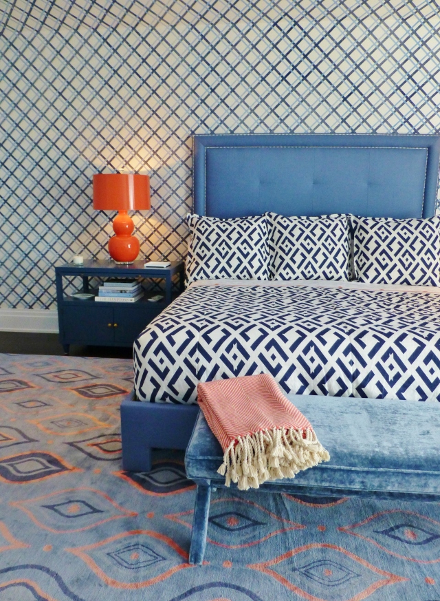 The bed features a solid blue upholstered headboard with double nailhead trim.  The bed, painted nightstands, and bold orange lamps provide a place for your eye to rest.