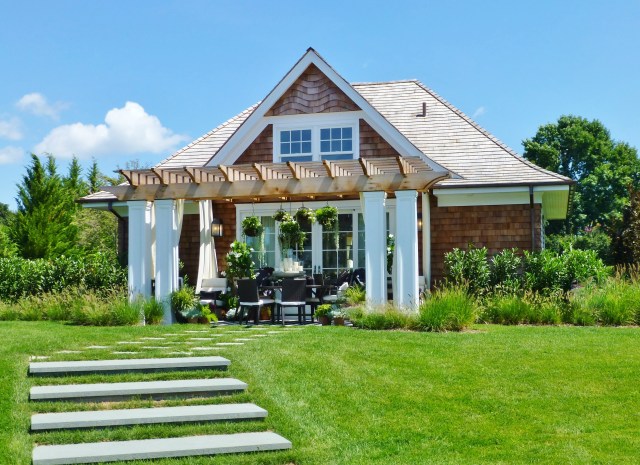 The opposite side of the pool house/guest cottage features a nearly identical terrace.  This one features a dining table and chairs, but has the same elegant black and white color scheme and elaborate hanging baskets.  Notice the chunky square white columns at the corners of the pergola.  The designer also placed white outdoor grommet draperies inside the pergola, to soften the look.