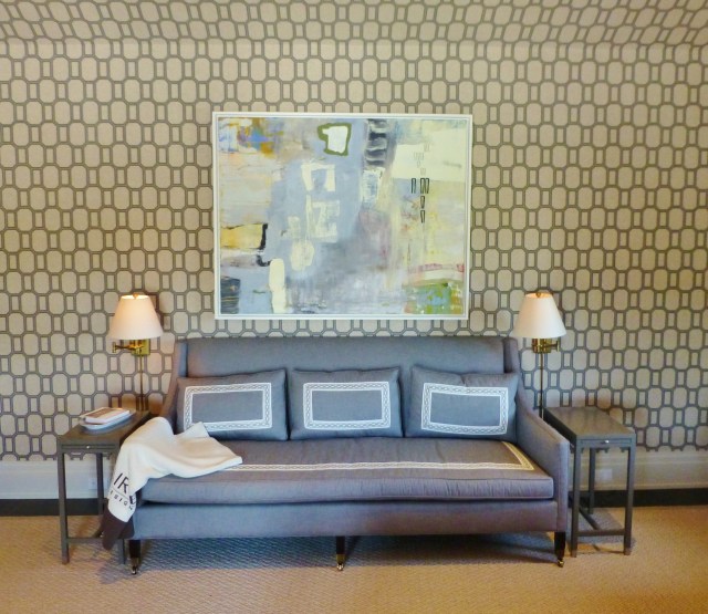 To the left of the bed (on the same wall as the entry door) is a gorgeous sofa upholstered in the same grey as the shades and bedhangings and using the same beautiful tape on the seat and pillows. A large piece of abstract artwork hangs above the sofa, flanked by swing arm lamps for reading. The side tables are very small and tailored, echoing the lines of the streamlined sofa.