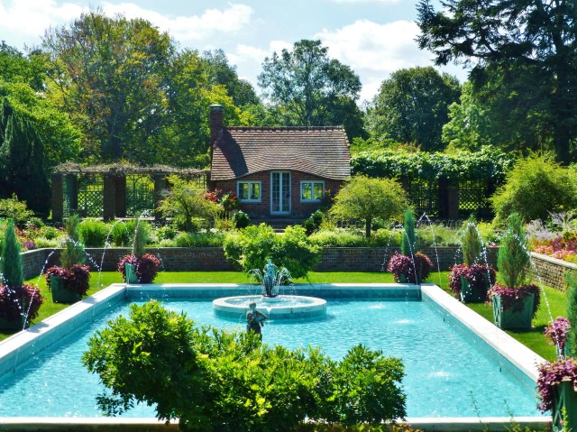 On closer inspection you will notice the adorable English Cottage, pool house at the far end of the pool. You can also see the amazing statuary placed amidst the pool fountains.   Large square painted wood planters frame out the pool with evergreens and trailing vines, which echo the color of the adorable red brick cottage.